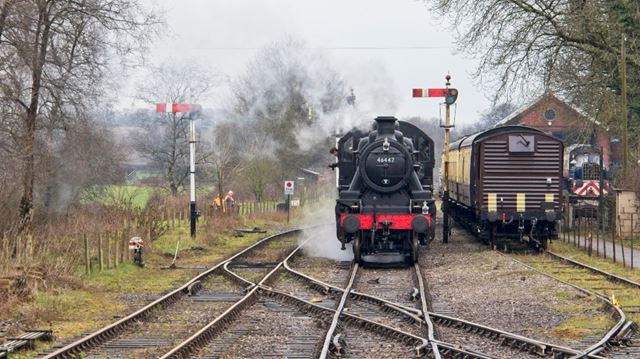 Ivatt locomotive engine 46447 steaming past brown carriages with railway tracks crossing in the foreground